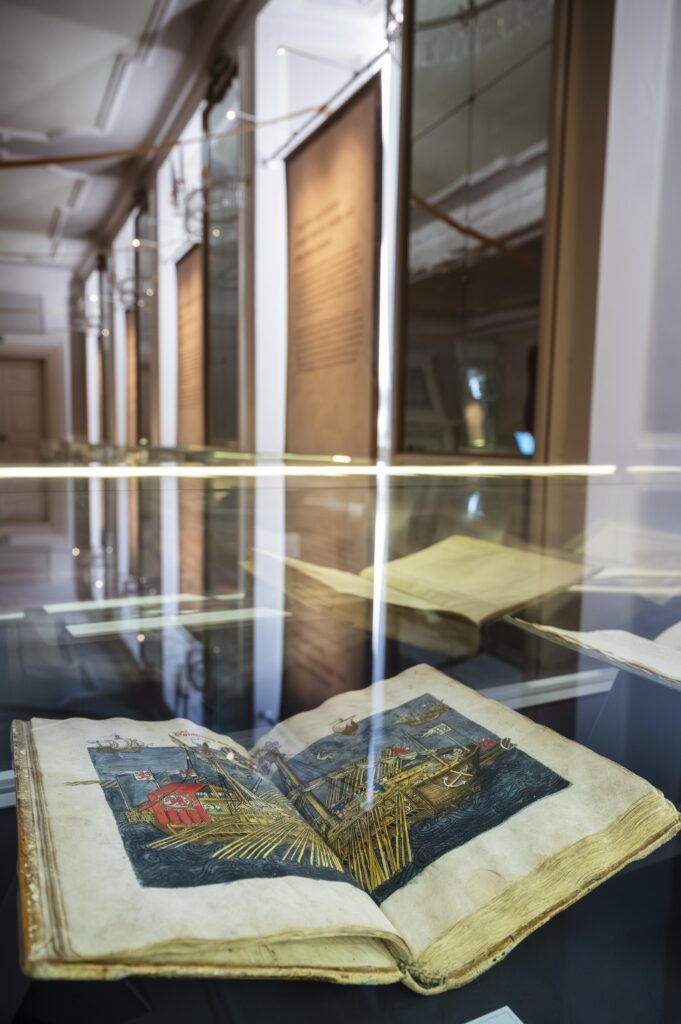Vitrine mit einem aufgeschlagenen Buch, das ein Pilgerschiff zeigt. Im Hintergrund Banner mit Ausstellungstexten